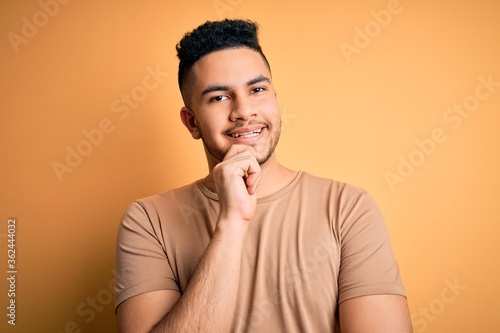 Young handsome man wearing casual t-shirt standing over isolated yellow background looking confident at the camera with smile with crossed arms and hand raised on chin. Thinking positive. © Krakenimages.com