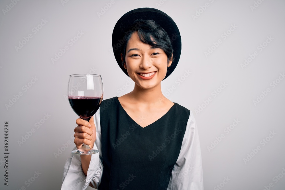 Young beautiful asian sommelier girl drinking glass of red wine over isolated white background with a happy face standing and smiling with a confident smile showing teeth Young beautiful asian sommelier girl drinking glass of red wine over isolated white background with a happy face standing and smiling with a confident smile showing teeth