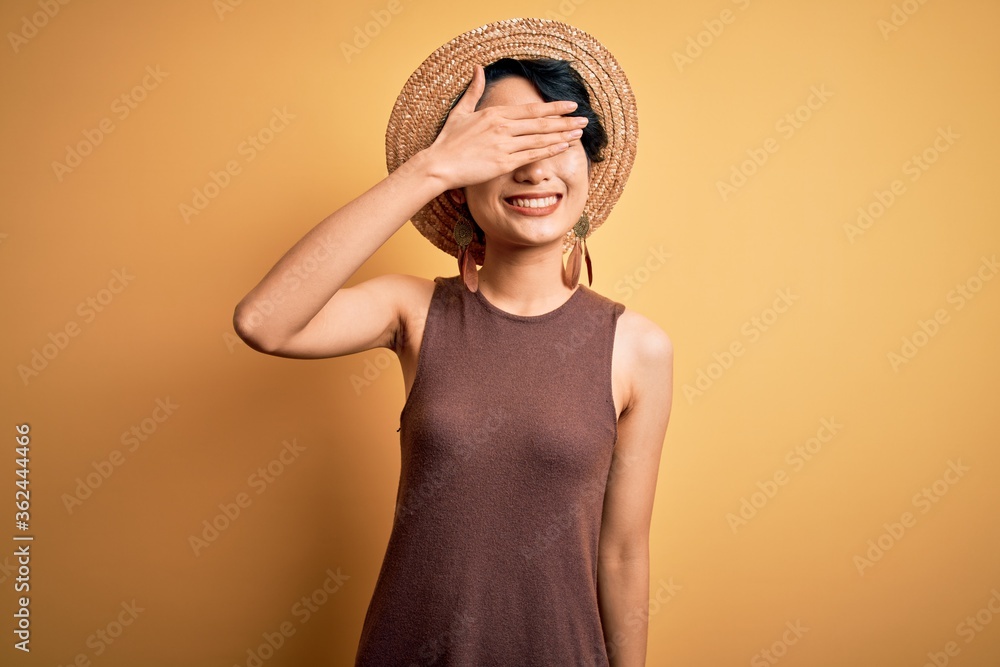 Young beautiful asian girl wearing casual t-shirt and hat over isolated yellow background smiling and laughing with hand on face covering eyes for surprise. Blind concept. Young beautiful asian girl wearing casual t-shirt and hat over isolated yellow background smiling and laughing with hand on face covering eyes for surprise. Blind concept.