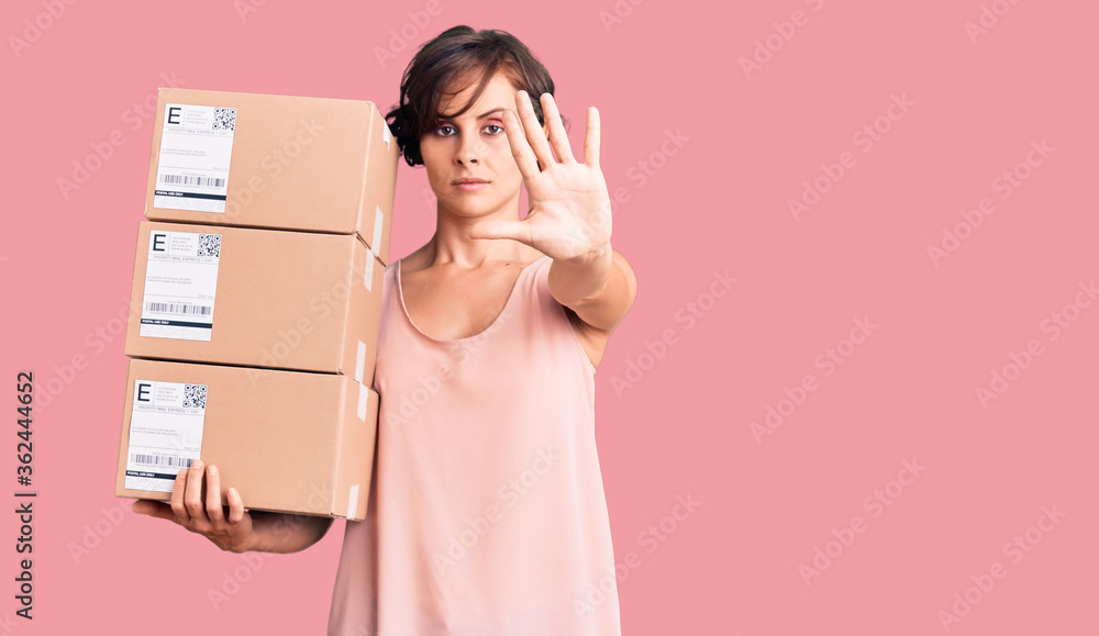 Beautiful young woman with short hair holding delivery packages with open hand doing stop sign with serious and confident expression, defense gesture Beautiful young woman with short hair holding delivery packages with open hand doing stop sign with serious and confident expression, defense gesture
