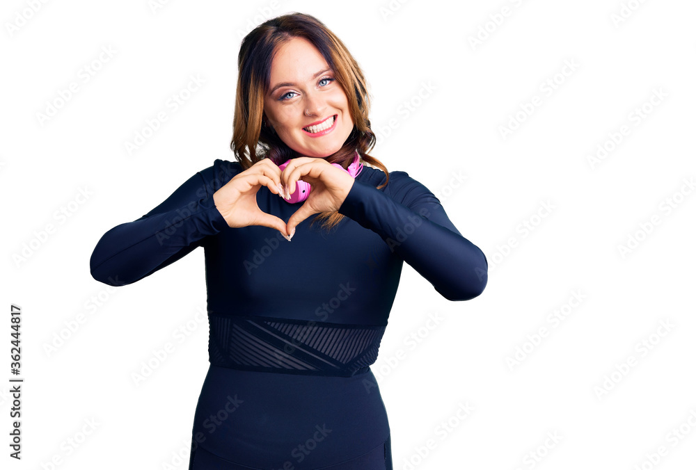 Young beautiful caucasian woman wearing gym clothes and using headphones smiling in love showing heart symbol and shape with hands. romantic concept. Young beautiful caucasian woman wearing gym clothes and using headphones smiling in love showing heart symbol and shape with hands. romantic concept.