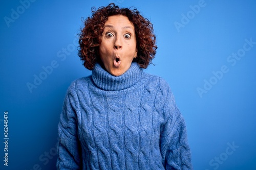 Middle age beautiful curly hair woman wearing casual turtleneck sweater over blue background afraid and shocked with surprise expression, fear and excited face.