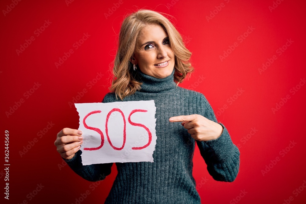 Middle age beautiful blonde woman holding paper with sos message over red bakground very happy pointing with hand and finger Middle age beautiful blonde woman holding paper with sos message over red bakground very happy pointing with hand and finger