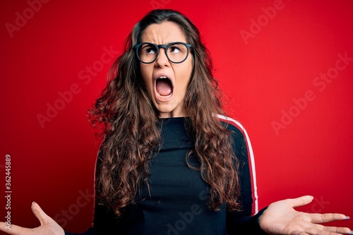 Young beautiful woman with curly hair wearing sweater and glasses over red background crazy and mad shouting and yelling with aggressive expression and arms raised. Frustration concept.