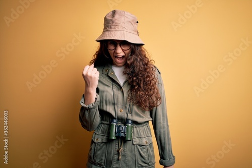 Young beautiful tourist woman on vacation wearing explorer hat and binoculars angry and mad raising fist frustrated and furious while shouting with anger. Rage and aggressive concept. © Krakenimages.com