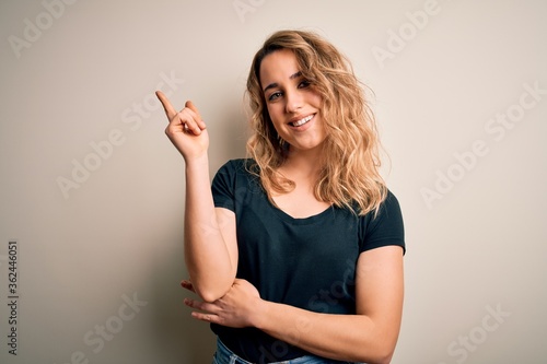 Young beautiful blonde woman wearing casual t-shirt standing over isolated white background with a big smile on face, pointing with hand and finger to the side looking at the camera.