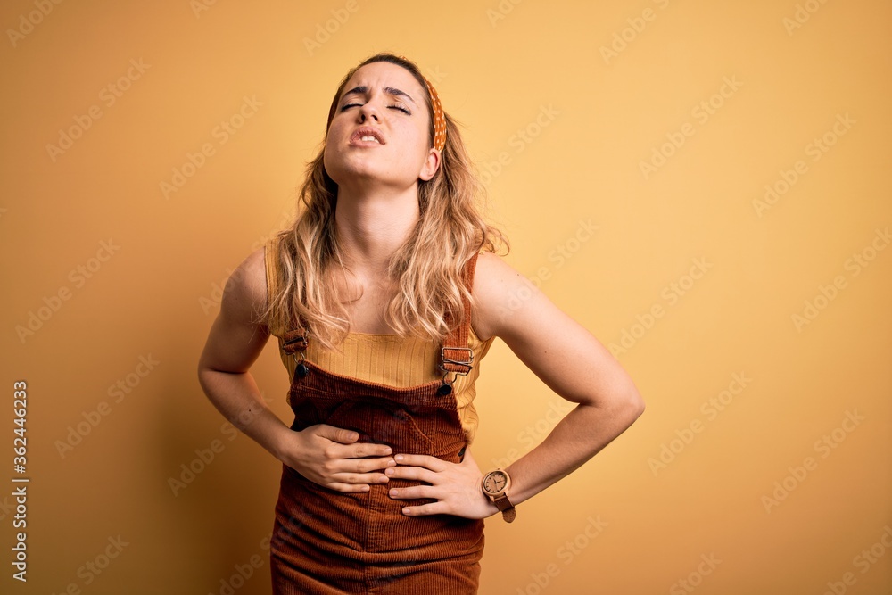 Young beautiful blonde woman wearing overalls and diadem standing over yellow background with hand on stomach because indigestion, painful illness feeling unwell. Ache concept. Young beautiful blonde woman wearing overalls and diadem standing over yellow background with hand on stomach because indigestion, painful illness feeling unwell. Ache concept.