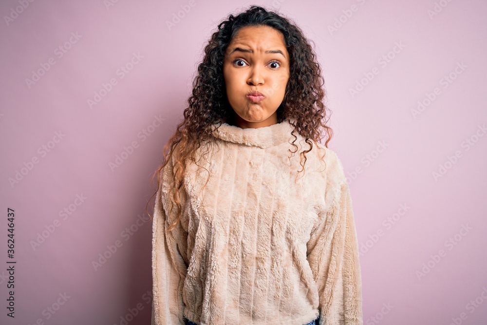 Young beautiful woman with curly hair wearing casual sweater standing over pink background puffing cheeks with funny face. Mouth inflated with air, crazy expression. Young beautiful woman with curly hair wearing casual sweater standing over pink background puffing cheeks with funny face. Mouth inflated with air, crazy expression.