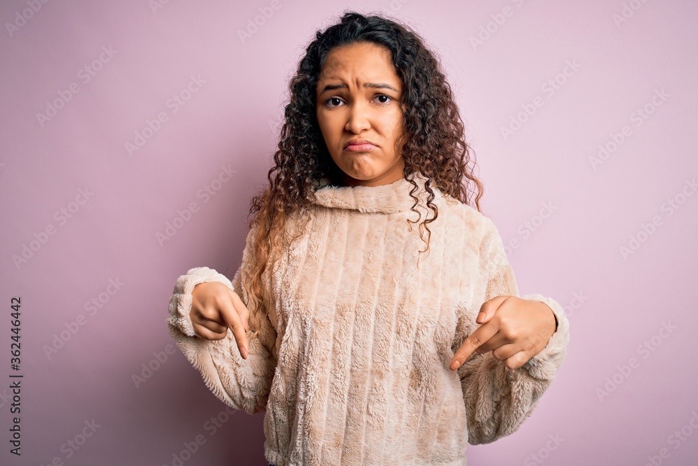 Young beautiful woman with curly hair wearing casual sweater standing over pink background Pointing down looking sad and upset, indicating direction with fingers, unhappy and depressed. Young beautiful woman with curly hair wearing casual sweater standing over pink background Pointing down looking sad and upset, indicating direction with fingers, unhappy and depressed.