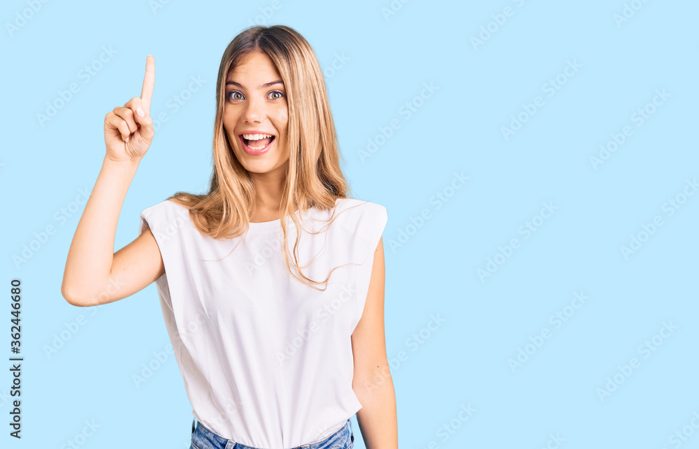 Beautiful caucasian woman with blonde hair wearing casual white tshirt pointing finger up with successful idea. exited and happy. number one.