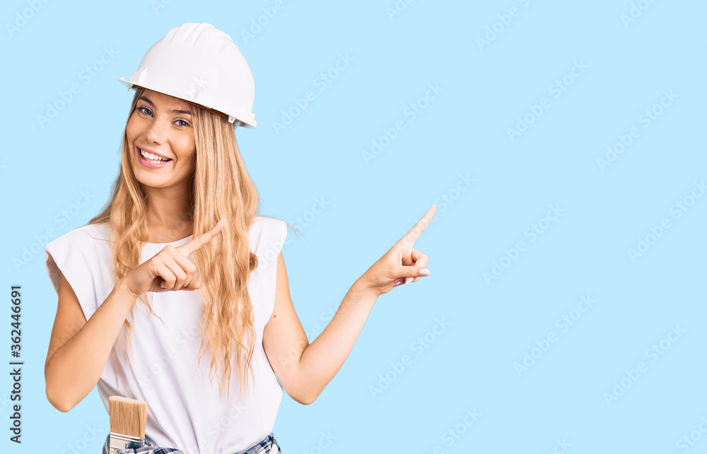 Beautiful caucasian woman with blonde hair wearing hardhat and painter clothes smiling and looking at the camera pointing with two hands and fingers to the side. Beautiful caucasian woman with blonde hair wearing hardhat and painter clothes smiling and looking at the camera pointing with two hands and fingers to the side.
