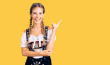 Beautiful caucasian woman with blonde hair wearing octoberfest traditional clothes with a big smile on face, pointing with hand and finger to the side looking at the camera.