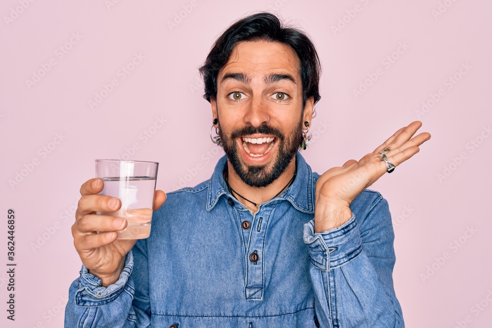 Young handsome hispanic bohemian hipster man with bear drinking a fresh glass of water very happy and excited, winner expression celebrating victory screaming with big smile and raised hands Young handsome hispanic bohemian hipster man with bear drinking a fresh glass of water very happy and excited, winner expression celebrating victory screaming with big smile and raised hands