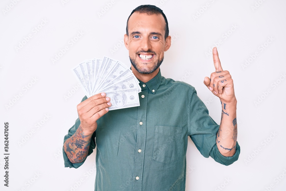Young handsome man with tattoo holding dollars surprised with an idea or question pointing finger with happy face, number one Young handsome man with tattoo holding dollars surprised with an idea or question pointing finger with happy face, number one