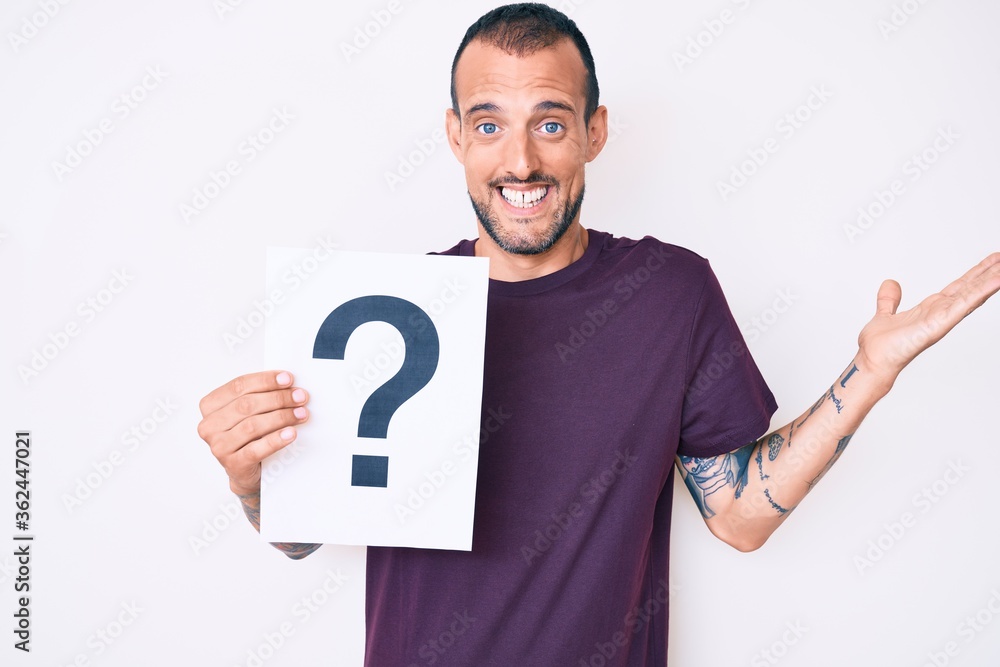 Young handsome man with tattoo holding question mark celebrating victory with happy smile and winner expression with raised hands Young handsome man with tattoo holding question mark celebrating victory with happy smile and winner expression with raised hands