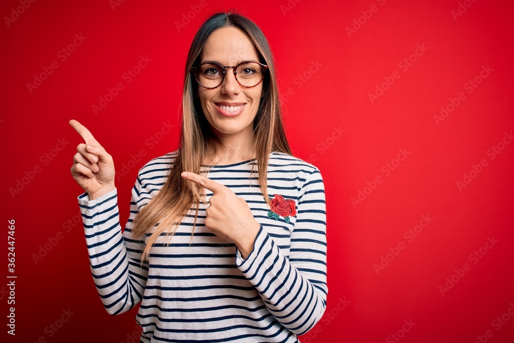 Young beautiful blonde woman with blue eyes wearing glasses standing over red background smiling and looking at the camera pointing with two hands and fingers to the side. Young beautiful blonde woman with blue eyes wearing glasses standing over red background smiling and looking at the camera pointing with two hands and fingers to the side.
