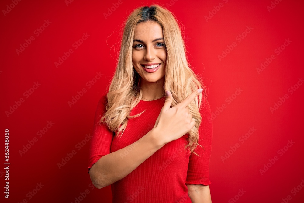 Young beautiful blonde woman wearing casual t-shirt standing over isolated red background cheerful with a smile on face pointing with hand and finger up to the side with happy and natural expression Young beautiful blonde woman wearing casual t-shirt standing over isolated red background cheerful with a smile on face pointing with hand and finger up to the side with happy and natural expression