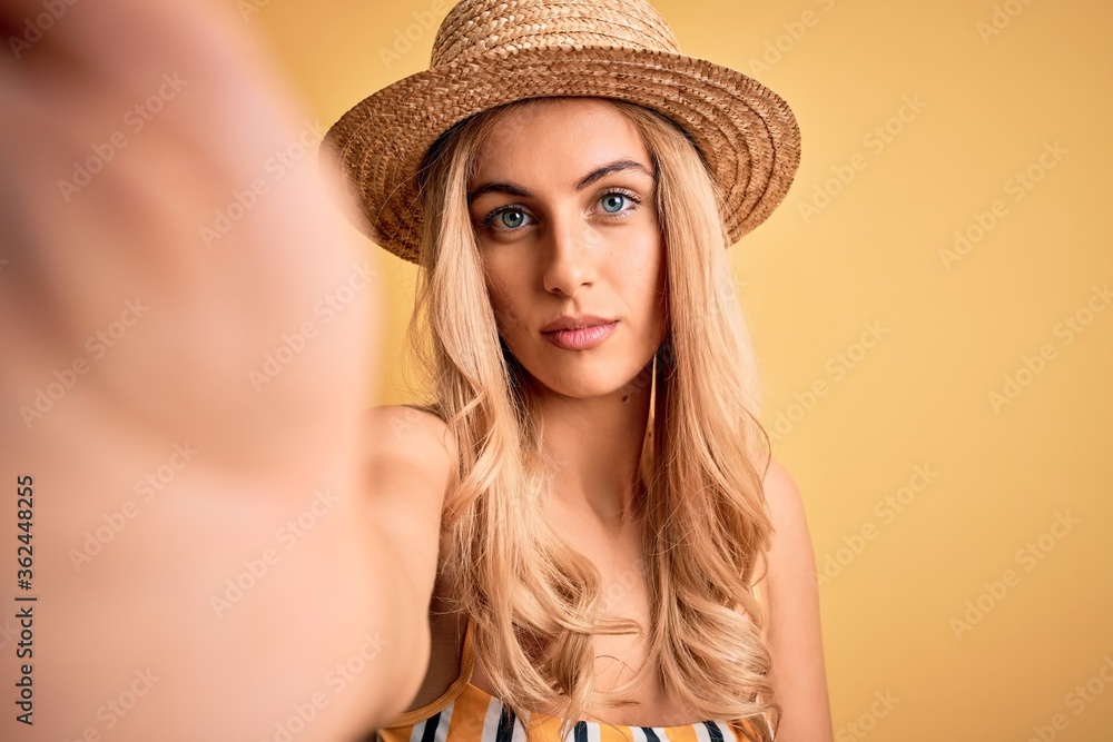 Young beautiful blonde woman on vacation wearing bikini and hat making selfie by camera with a confident expression on smart face thinking serious Young beautiful blonde woman on vacation wearing bikini and hat making selfie by camera with a confident expression on smart face thinking serious