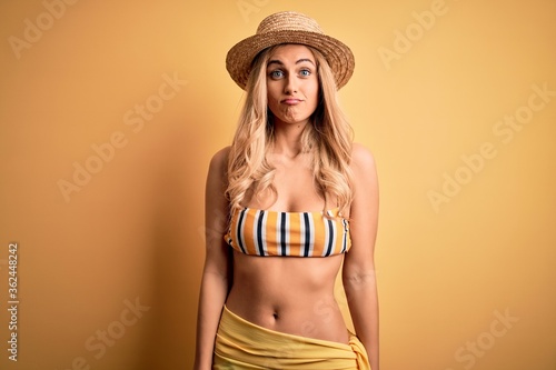 Young beautiful blonde woman on vacation wearing bikini and hat over yellow background puffing cheeks with funny face. Mouth inflated with air  crazy expression.