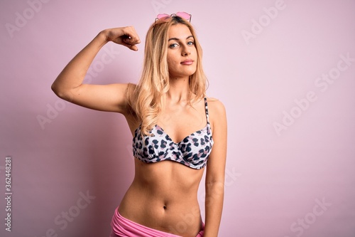 Young beautiful blonde woman on vacation wearing bikini over isolated pink background Strong person showing arm muscle, confident and proud of power © Krakenimages.com