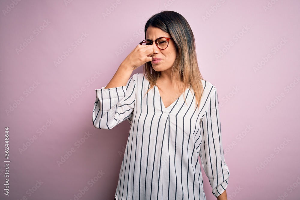 Young beautiful woman wearing casual striped t-shirt and glasses over pink background smelling something stinky and disgusting, intolerable smell, holding breath with fingers on nose. Bad smell Young beautiful woman wearing casual striped t-shirt and glasses over pink background smelling something stinky and disgusting, intolerable smell, holding breath with fingers on nose. Bad smell