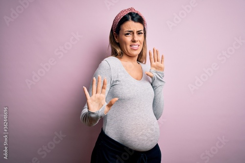 Young beautiful brunette woman pregnant expecting baby over isolated pink background disgusted expression, displeased and fearful doing disgust face because aversion reaction. With hands raised photo