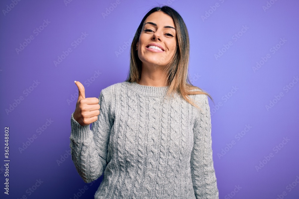 Young beautiful woman wearing casual sweater standing over isolated purple background doing happy thumbs up gesture with hand. Approving expression looking at the camera showing success.