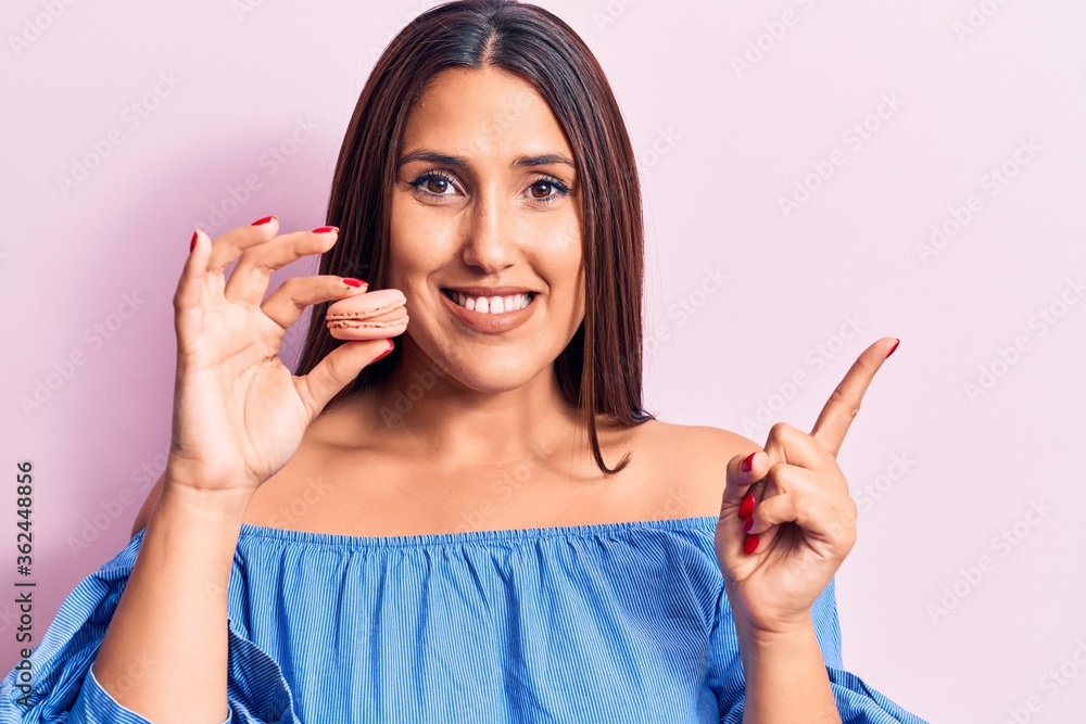 Young beautiful brunette woman holding macaron smiling happy pointing with hand and finger to the side Young beautiful brunette woman holding macaron smiling happy pointing with hand and finger to the side