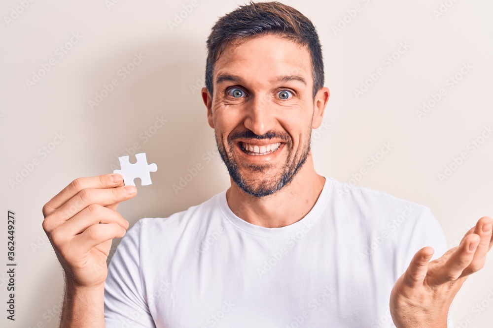 Young handsome man holding piece of puzzle standing over isolated white background celebrating achievement with happy smile and winner expression with raised hand Young handsome man holding piece of puzzle standing over isolated white background celebrating achievement with happy smile and winner expression with raised hand