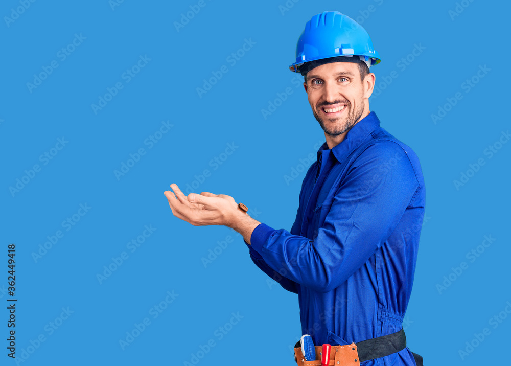 Young handsome man wearing worker uniform and hardhat inviting to enter smiling natural with open hand