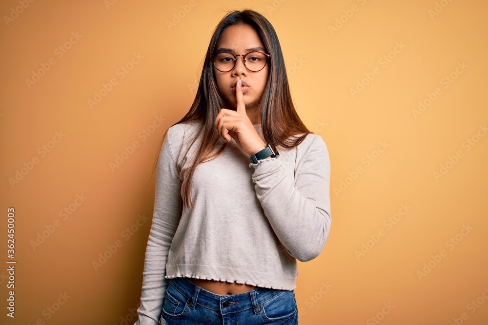 Young beautiful asian girl wearing casual sweater and glasses over yellow background asking to be quiet with finger on lips. Silence and secret concept. Young beautiful asian girl wearing casual sweater and glasses over yellow background asking to be quiet with finger on lips. Silence and secret concept.