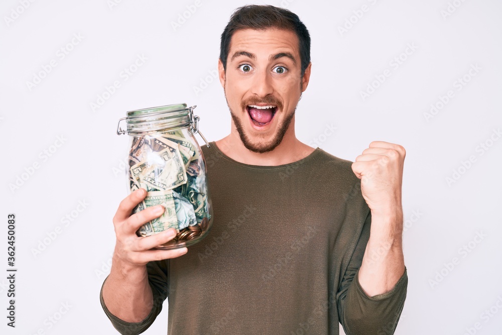 Young handsome man holding jar with savings screaming proud, celebrating victory and success very excited with raised arms Young handsome man holding jar with savings screaming proud, celebrating victory and success very excited with raised arms