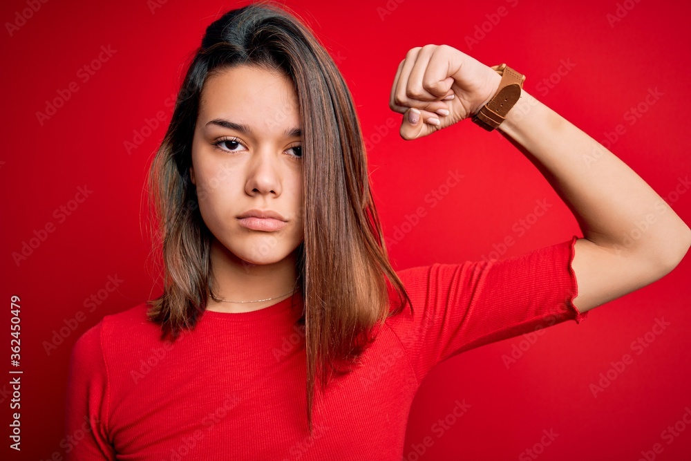 Young beautiful brunette girl wearing casual t-shirt over isolated red background Strong person showing arm muscle, confident and proud of power Young beautiful brunette girl wearing casual t-shirt over isolated red background Strong person showing arm muscle, confident and proud of power
