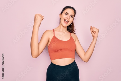 Young beautiful fitness woman wearing sport excersie clothes over pink background celebrating surprised and amazed for success with arms raised and open eyes. Winner concept.