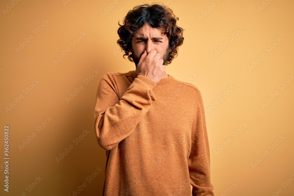 Young handsome man with beard wearing casual sweater standing over yellow background smelling something stinky and disgusting, intolerable smell, holding breath with fingers on nose. Bad smell Young handsome man with beard wearing casual sweater standing over yellow background smelling something stinky and disgusting, intolerable smell, holding breath with fingers on nose. Bad smell
