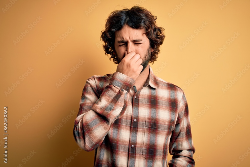 Young handsome man with beard wearing casual shirt standing over yellow background smelling something stinky and disgusting, intolerable smell, holding breath with fingers on nose. Bad smell Young handsome man with beard wearing casual shirt standing over yellow background smelling something stinky and disgusting, intolerable smell, holding breath with fingers on nose. Bad smell
