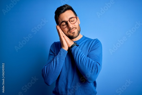 Young handsome man with beard wearing casual sweater and glasses over blue background sleeping tired dreaming and posing with hands together while smiling with closed eyes. © Krakenimages.com