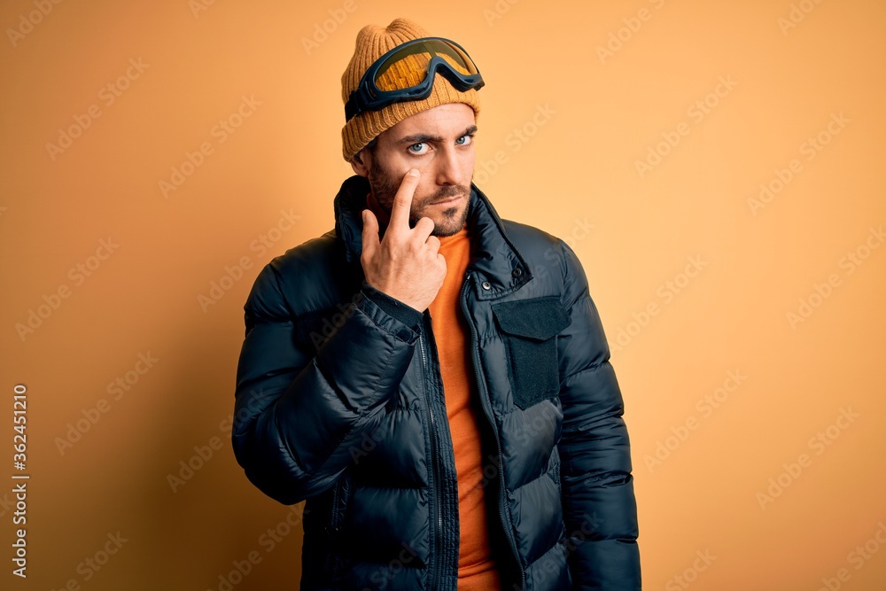Young handsome skier man with beard wearing snow sportswear and ski goggles Pointing to the eye watching you gesture, suspicious expression Young handsome skier man with beard wearing snow sportswear and ski goggles Pointing to the eye watching you gesture, suspicious expression