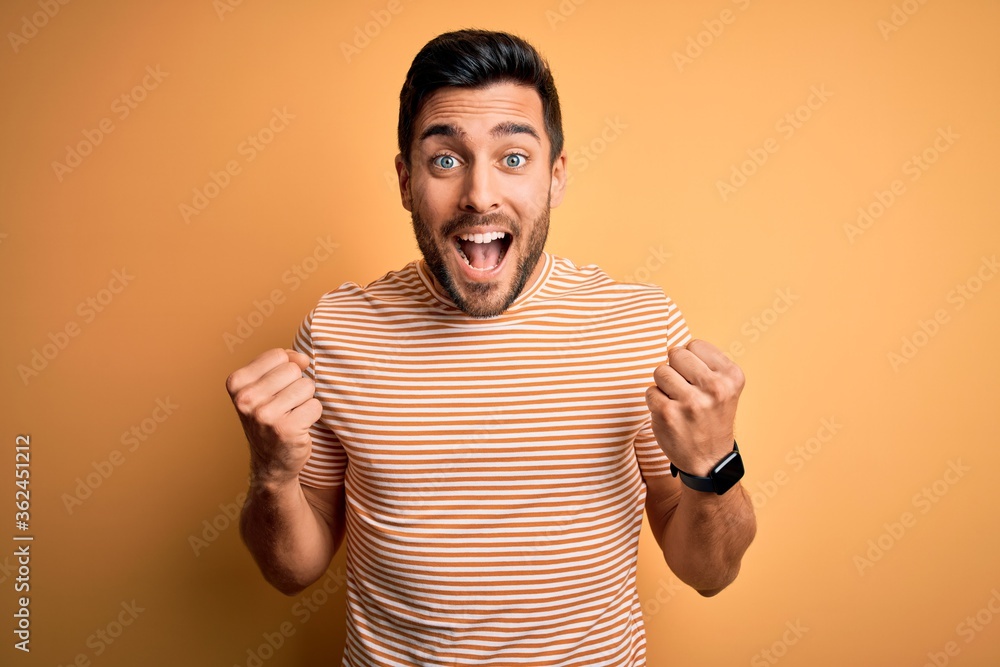 Young handsome man with beard wearing casual striped t-shirt over yellow background celebrating surprised and amazed for success with arms raised and open eyes. Winner concept. Young handsome man with beard wearing casual striped t-shirt over yellow background celebrating surprised and amazed for success with arms raised and open eyes. Winner concept.