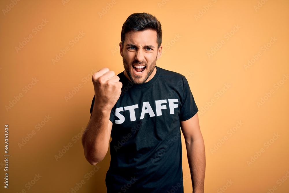 Young handsome worker man with beard wearing staff uniform t-shirt over yellow background annoyed and frustrated shouting with anger, crazy and yelling with raised hand, anger concept Young handsome worker man with beard wearing staff uniform t-shirt over yellow background annoyed and frustrated shouting with anger, crazy and yelling with raised hand, anger concept