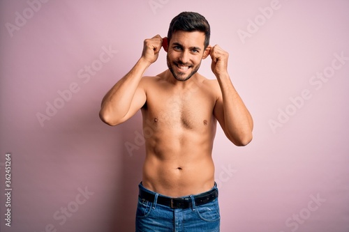 Young handsome strong man with beard shirtless standing over isolated pink background Smiling pulling ears with fingers, funny gesture. Audition problem