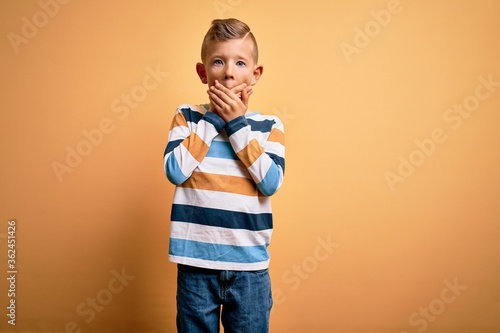 Young little caucasian kid with blue eyes wearing colorful striped shirt over yellow background shocked covering mouth with hands for mistake. Secret concept.