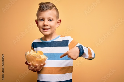 Young little caucasian kid eating unheatlhy potatoes crisps chips over yellow background with surprise face pointing finger to himself photo