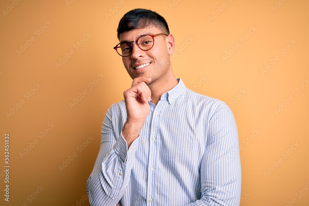 Young handsome hispanic business man wearing nerd glasses over yellow background looking confident at the camera with smile with crossed arms and hand raised on chin. Thinking positive. Young handsome hispanic business man wearing nerd glasses over yellow background looking confident at the camera with smile with crossed arms and hand raised on chin. Thinking positive.