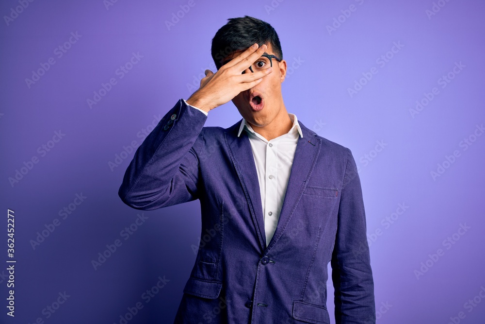 Young handsome business man wearing jacket and glasses over isolated purple background peeking in shock covering face and eyes with hand, looking through fingers with embarrassed expression. Young handsome business man wearing jacket and glasses over isolated purple background peeking in shock covering face and eyes with hand, looking through fingers with embarrassed expression.