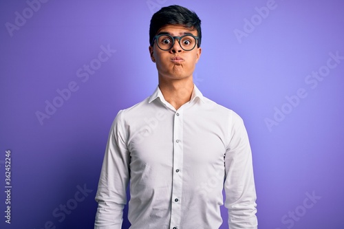 Young handsome business man wearing shirt and glasses over isolated purple background puffing cheeks with funny face. Mouth inflated with air, crazy expression.