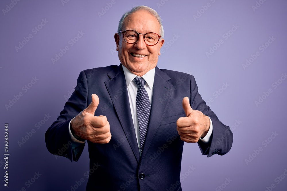 Grey haired senior business man wearing glasses and elegant suit and tie over purple background success sign doing positive gesture with hand, thumbs up smiling and happy. Cheerful expression. Grey haired senior business man wearing glasses and elegant suit and tie over purple background success sign doing positive gesture with hand, thumbs up smiling and happy. Cheerful expression.