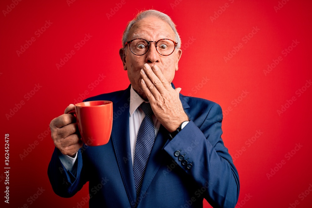 Senior grey haired business man drinking a hot cup of coffee over red background cover mouth with hand shocked with shame for mistake, expression of fear, scared in silence, secret concept Senior grey haired business man drinking a hot cup of coffee over red background cover mouth with hand shocked with shame for mistake, expression of fear, scared in silence, secret concept