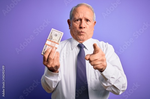 Senior grey haired man holding bunch of fifty dollars banknotes over purple background pointing with finger to the camera and to you, hand sign, positive and confident gesture from the front