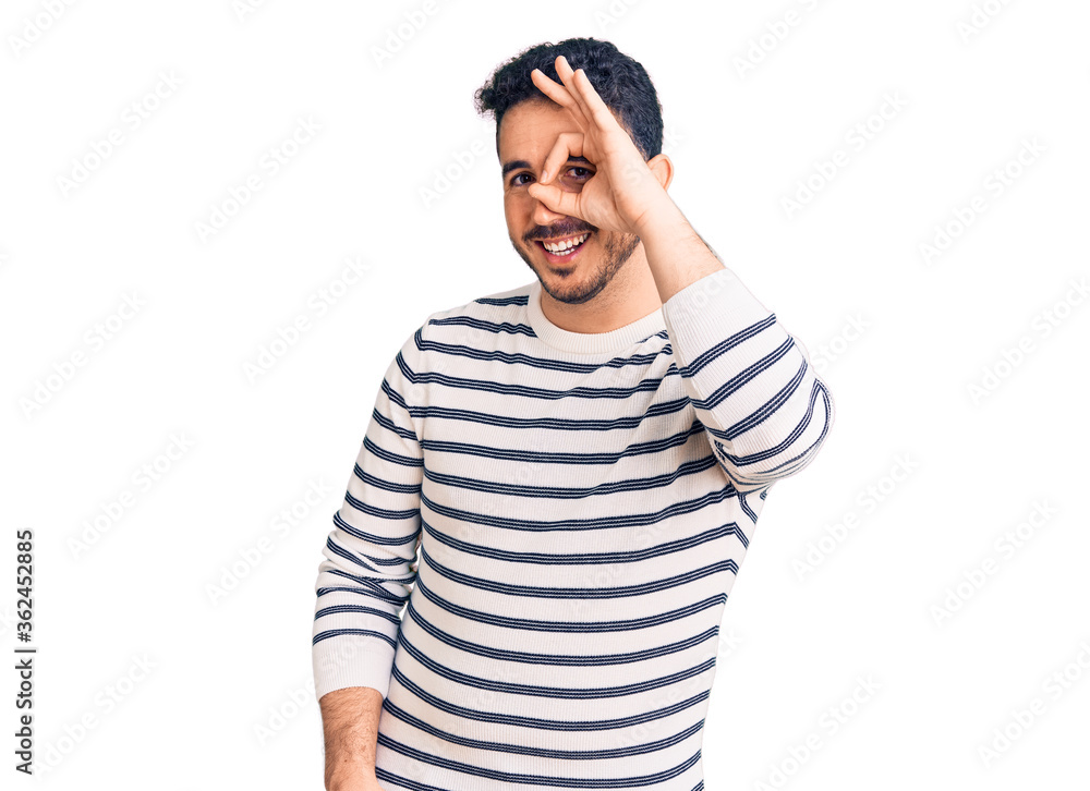 Young hispanic man wearing casual clothes doing ok gesture with hand smiling, eye looking through fingers with happy face. Young hispanic man wearing casual clothes doing ok gesture with hand smiling, eye looking through fingers with happy face.